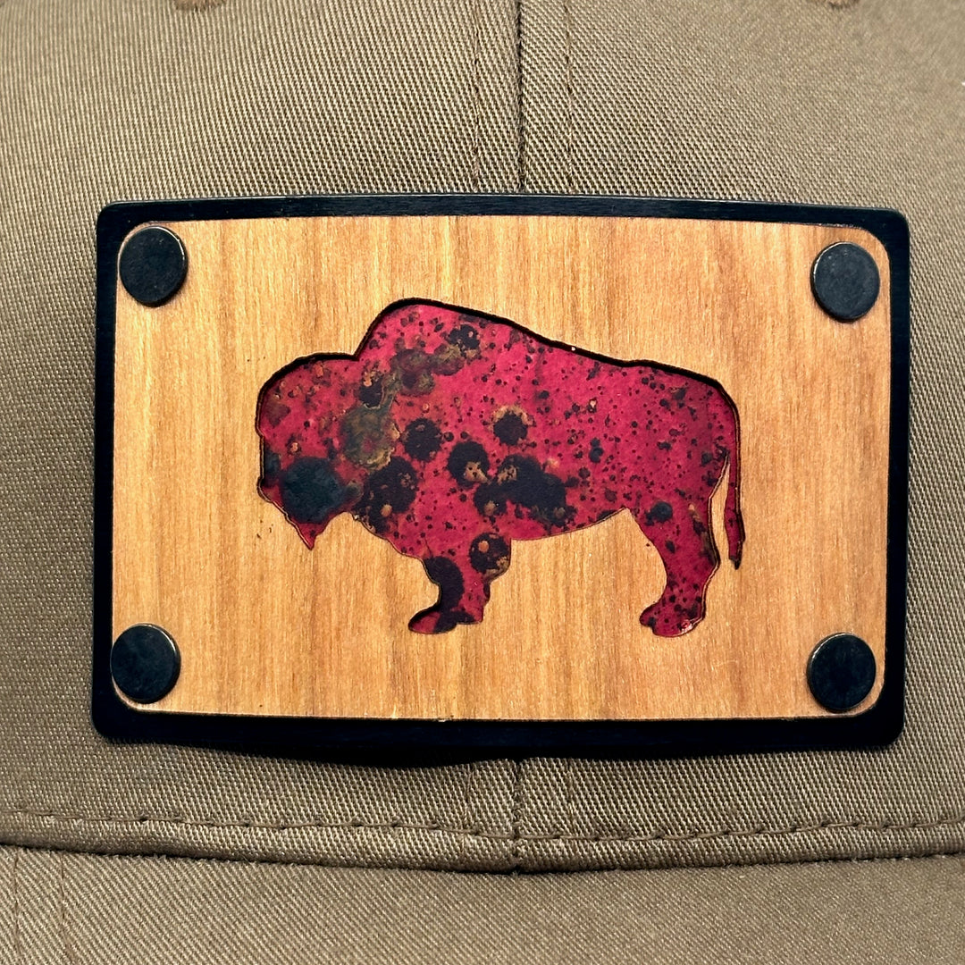 A detail of Cherry Wood & Red Copper Bison Coyote Brown & Black Trucker Hat, featuring a rectangular real cherry wood veneer patch with cut-out bison silhouette inlayed with red copper, laminated to a black anodized aluminum plate, and riveted to a coyote brown and black mesh Flexfit 110M trucker hat—handmade in Montana, USA by Last Best Supply Co.