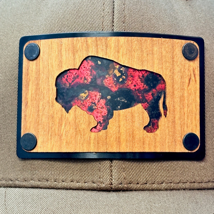 A detail of Cherry Wood & Red Copper Bison Coyote Brown & Black Trucker Hat, featuring a rectangular real cherry wood veneer patch with cut-out bison silhouette inlayed with red copper, laminated to a black anodized aluminum plate, and riveted to a coyote brown and black mesh Flexfit 110M trucker hat—handmade in Montana, USA by Last Best Supply Co.