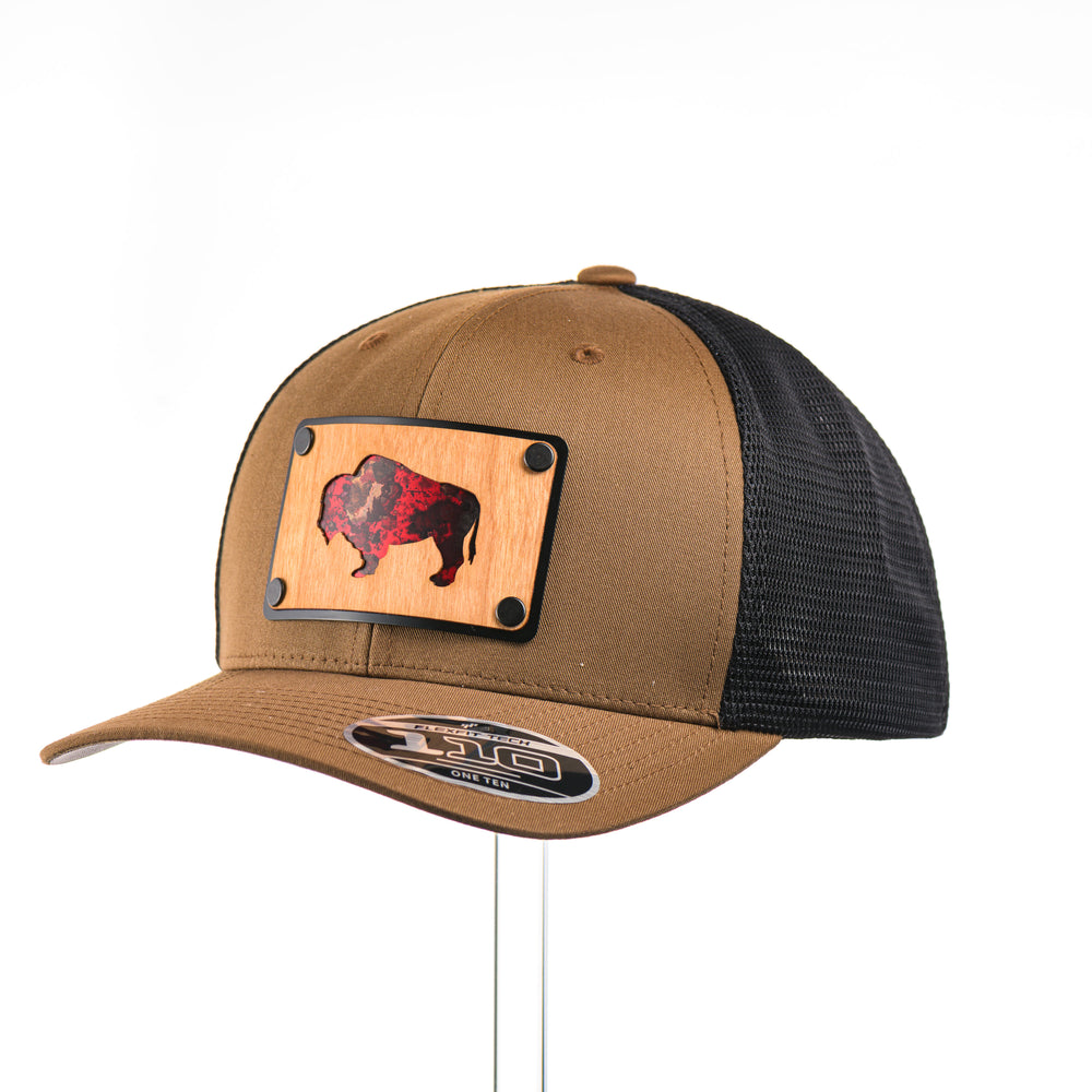 3/4 view of a Cherry Wood & Red Copper Bison Coyote Brown & Black Trucker Hat, featuring a rectangular real cherry wood veneer patch with cut-out bison silhouette inlayed with red copper, laminated to a black anodized aluminum plate, and riveted to a coyote brown and black mesh Flexfit 110M trucker hat—handmade in Montana, USA by Last Best Supply Co.