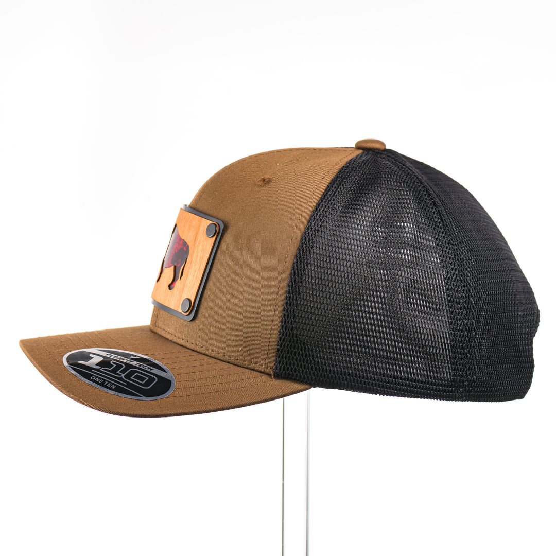 A side view of a Cherry Wood & Red Copper Bison Coyote Brown & Black Trucker Hat, featuring a rectangular real cherry wood veneer patch with cut-out bison silhouette inlayed with red copper, laminated to a black anodized aluminum plate, and riveted to a coyote brown and black mesh Flexfit 110M trucker hat—handmade in Montana, USA by Last Best Supply Co.