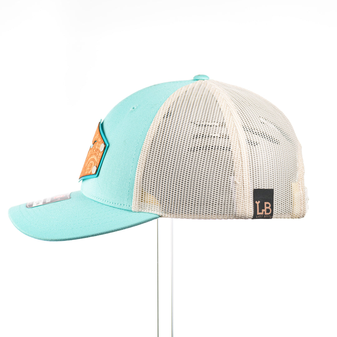 The side of a Cherry Wood Steer Skull w/ Paisley Teal & Cream Trucker Hat, featuring a hexagonal real cherry wood veneer patch with cut-out steer skull silhouette surrounded by etched paisley design, laminated to a hexagonal teal anodized aluminum plate, and riveted to a light teal and cream Richardson 115 low-profile trucker hat—handmade in Montana, USA by Last Best Supply Co.