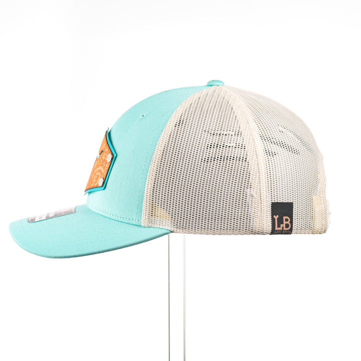 The side of a Cherry Wood Steer Skull w/ Paisley Teal & Cream Trucker Hat, featuring a hexagonal real cherry wood veneer patch with cut-out steer skull silhouette surrounded by etched paisley design, laminated to a hexagonal teal anodized aluminum plate, and riveted to a light teal and cream Richardson 115 low-profile trucker hat—handmade in Montana, USA by Last Best Supply Co.