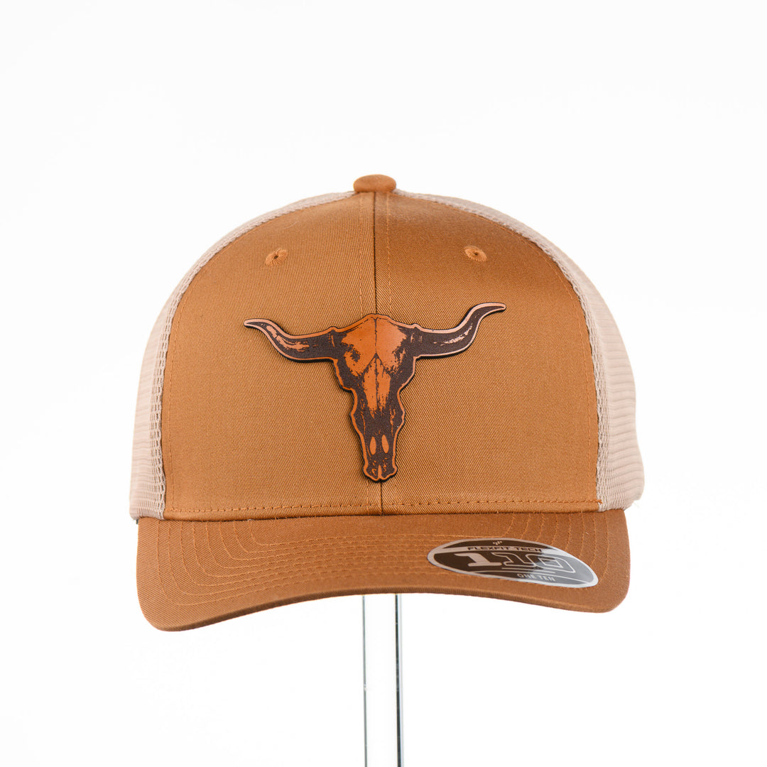 Etched Leather Bull Skull Patch Flexfit Trucker Hat, featuring a real leather patch cut into the shape of a steer skull and etched with detail, securely attached to a caramel and khaki trucker hat—handmade in Montana, USA by Last Best Supply Co.