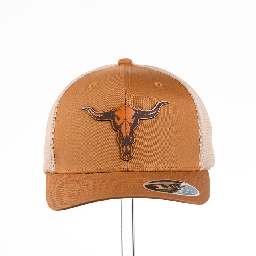 Etched Leather Bull Skull Patch Flexfit Trucker Hat, featuring a real leather patch cut into the shape of a steer skull and etched with detail, securely attached to a caramel and khaki trucker hat—handmade in Montana, USA by Last Best Supply Co.