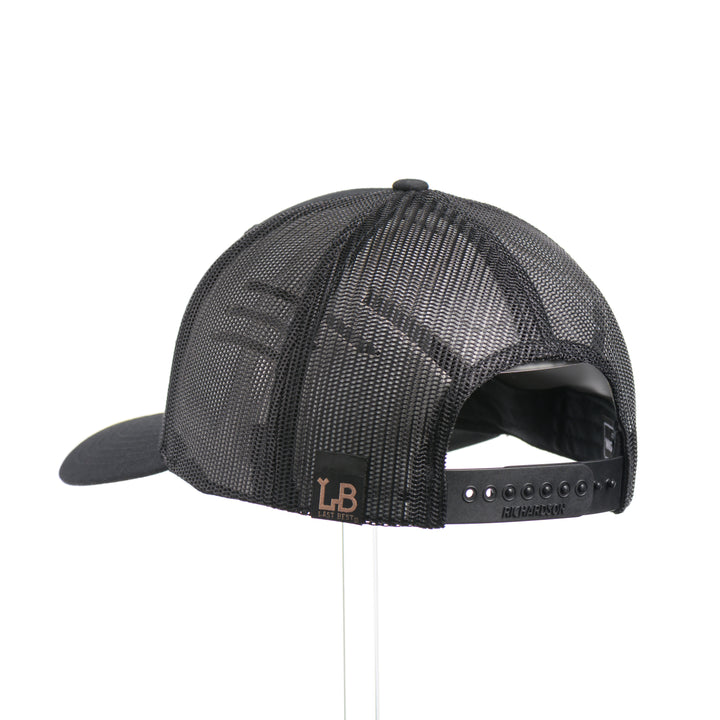 The back of an Etched Mahogany Camping Triangle Patch Trucker Hat, featuring a real mahogany wood veneer patch precision-cut into a hexagon with a forest scene cut-out centered around a camping tent, laminated to a hexagonal green anodized aluminum plate, and riveted to an all-black Richardson 112 trucker hat—handmade in Montana, USA by Last Best Supply Co.