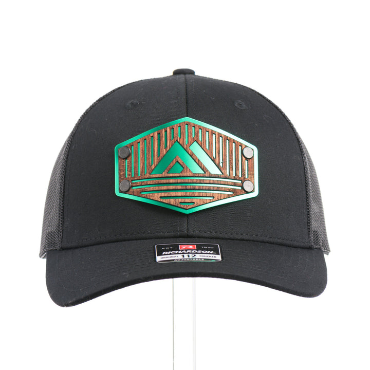 The front of an Etched Mahogany Camping Triangle Patch Trucker Hat, featuring a real mahogany wood veneer patch precision-cut into a hexagon with a forest scene cut-out centered around a camping tent, laminated to a hexagonal green anodized aluminum plate, and riveted to an all-black Richardson 112 trucker hat—handmade in Montana, USA by Last Best Supply Co.