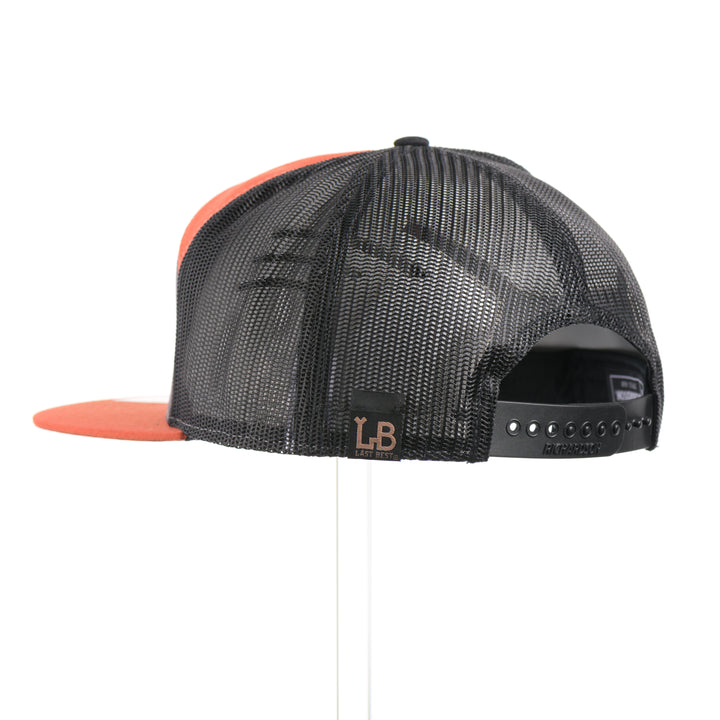 The back of a Mahogany Wood Bison Sunrise Flat Bill Hat, featuring a real mahogany wood veneer patch precision-cut into a hexagon, etched with a rising sun with a cut-out silhouette of a bison, laminated to a hexagonal orange anodized aluminum plate, and riveted to a burnt orange, charcoal gray, and black mesh Richardson 168 flat bill trucker hat—handmade in Montana, USA by Last Best Supply Co.