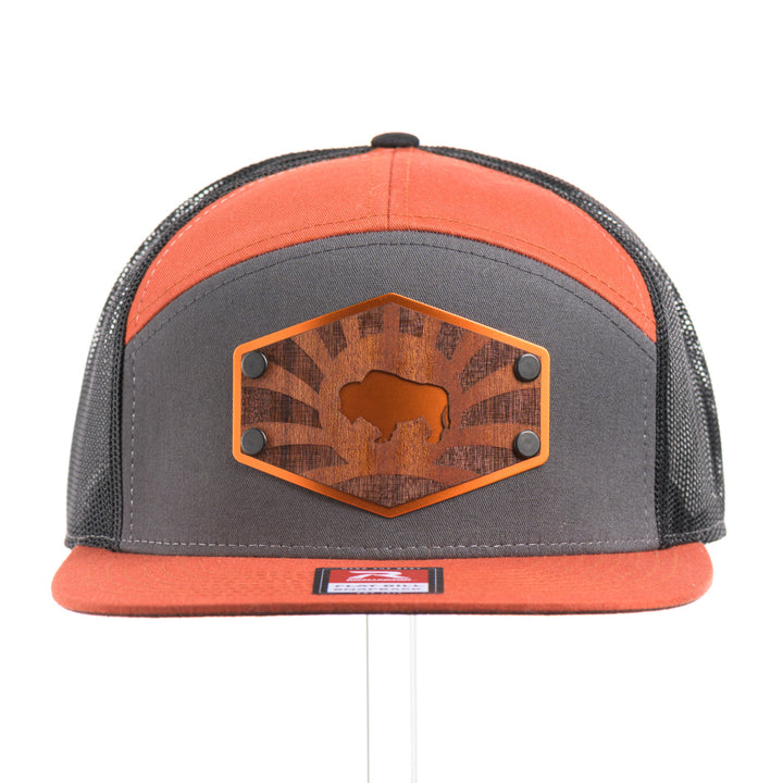 The front of a Mahogany Wood Bison Sunrise Flat Bill Hat, featuring a real mahogany wood veneer patch precision-cut into a hexagon, etched with a rising sun with a cut-out silhouette of a bison, laminated to a hexagonal orange anodized aluminum plate, and riveted to a burnt orange, charcoal gray, and black mesh Richardson 168 flat bill trucker hat—handmade in Montana, USA by Last Best Supply Co.