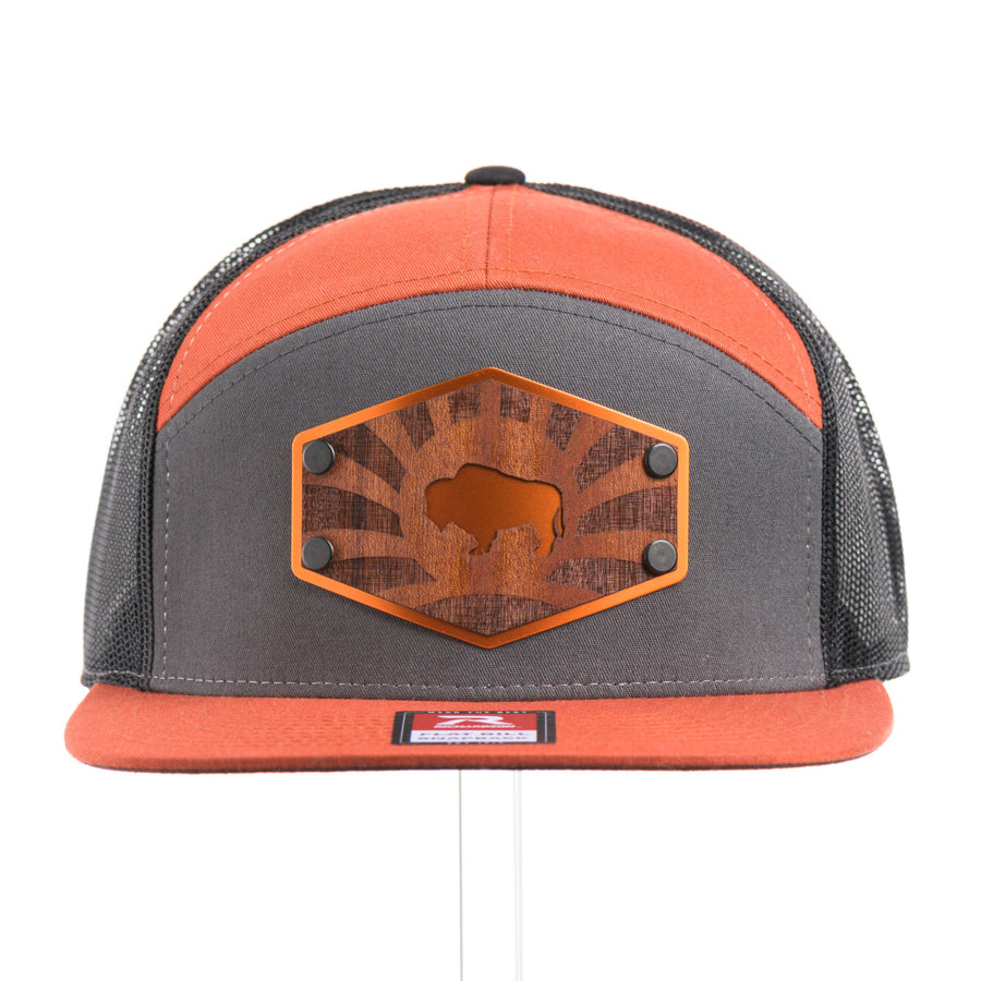 The front of a Mahogany Wood Bison Sunrise Flat Bill Hat, featuring a real mahogany wood veneer patch precision-cut into a hexagon, etched with a rising sun with a cut-out silhouette of a bison, laminated to a hexagonal orange anodized aluminum plate, and riveted to a burnt orange, charcoal gray, and black mesh Richardson 168 flat bill trucker hat—handmade in Montana, USA by Last Best Supply Co.