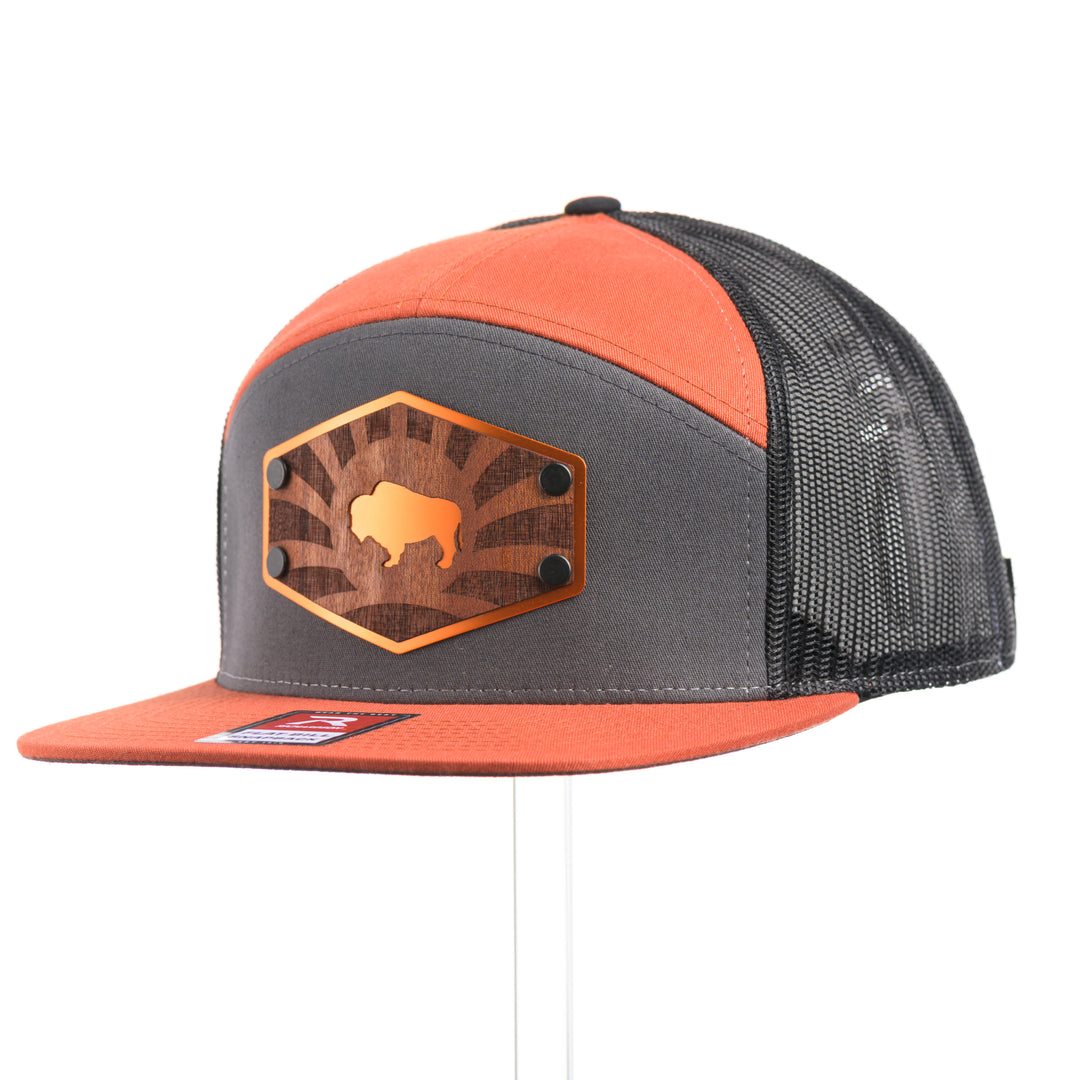 3/4 view of a Mahogany Wood Bison Sunrise Flat Bill Hat, featuring a real mahogany wood veneer patch precision-cut into a hexagon, etched with a rising sun with a cut-out silhouette of a bison, laminated to a hexagonal orange anodized aluminum plate, and riveted to a burnt orange, charcoal gray, and black mesh Richardson 168 flat bill trucker hat—handmade in Montana, USA by Last Best Supply Co.