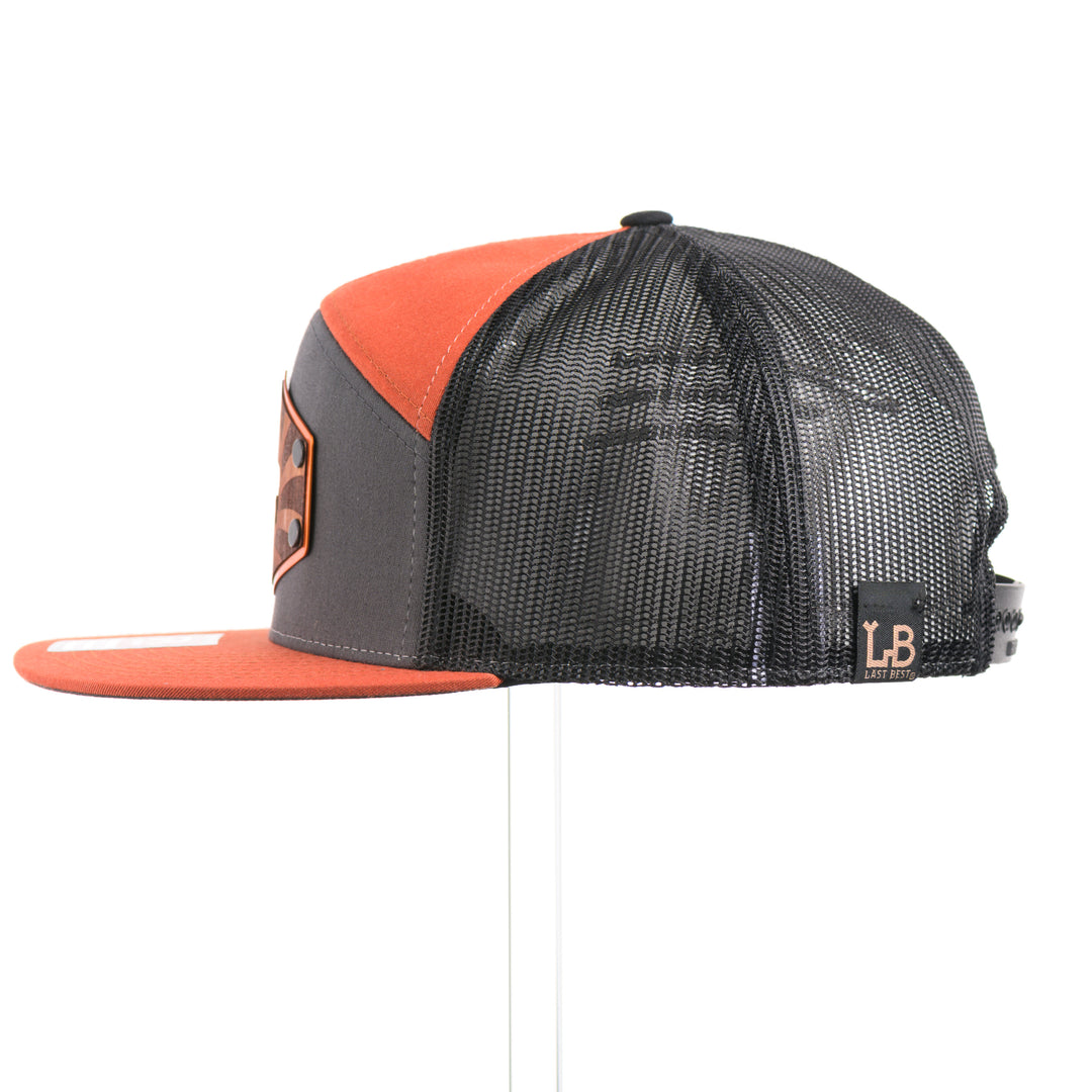 The side of a Mahogany Wood Bison Sunrise Flat Bill Hat, featuring a real mahogany wood veneer patch precision-cut into a hexagon, etched with a rising sun with a cut-out silhouette of a bison, laminated to a hexagonal orange anodized aluminum plate, and riveted to a burnt orange, charcoal gray, and black mesh Richardson 168 flat bill trucker hat—handmade in Montana, USA by Last Best Supply Co.