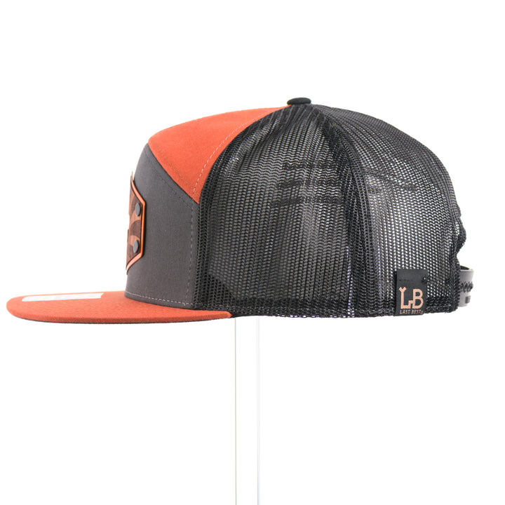 The side of a Mahogany Wood Bison Sunrise Flat Bill Hat, featuring a real mahogany wood veneer patch precision-cut into a hexagon, etched with a rising sun with a cut-out silhouette of a bison, laminated to a hexagonal orange anodized aluminum plate, and riveted to a burnt orange, charcoal gray, and black mesh Richardson 168 flat bill trucker hat—handmade in Montana, USA by Last Best Supply Co.