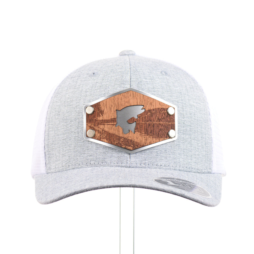 The front of a Mahogany Wood Jumping Trout Patch Flexfit Trucker Hat, featuring a real mahogany wood patch cut into the silhouette of trees, mounted on an anodized aluminum plate and secured with silver rivets to a heathered silver and white Flexfit trucker hat—handmade in Montana, USA by Last Best Supply Co.