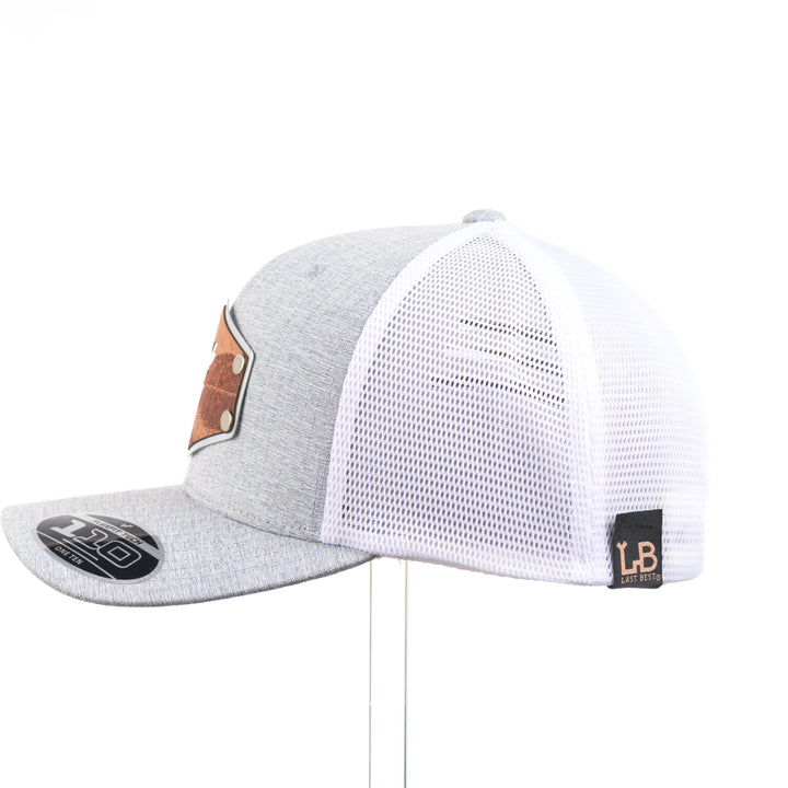 The side of a Mahogany Wood Jumping Trout Patch Flexfit Trucker Hat, featuring a real mahogany wood patch cut into the silhouette of trees, mounted on an anodized aluminum plate and secured with silver rivets to a heathered silver and white Flexfit trucker hat—handmade in Montana, USA by Last Best Supply Co.