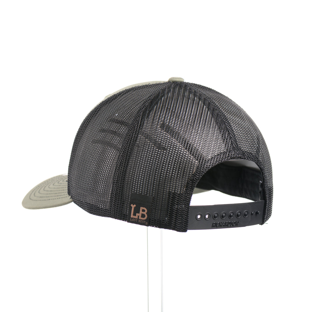 The back of a Walnut Wood Bubble Salmon Patch Trucker Hat, featuring a real walnut wood veneer patch precision-cut into a hexagon, etched with a river bubbles design with a cut-out silhouette of a salmon, laminated to a hexagonal black anodized aluminum plate, and riveted to a loden green and black mesh Richardson 112 trucker hat—handmade in Montana, USA by Last Best Supply Co.