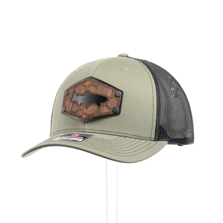 3/4 view of a Walnut Wood Bubble Salmon Patch Trucker Hat, featuring a real walnut wood veneer patch precision-cut into a hexagon, etched with a river bubbles design with a cut-out silhouette of a salmon, laminated to a hexagonal black anodized aluminum plate, and riveted to a loden green and black mesh Richardson 112 trucker hat—handmade in Montana, USA by Last Best Supply Co.
