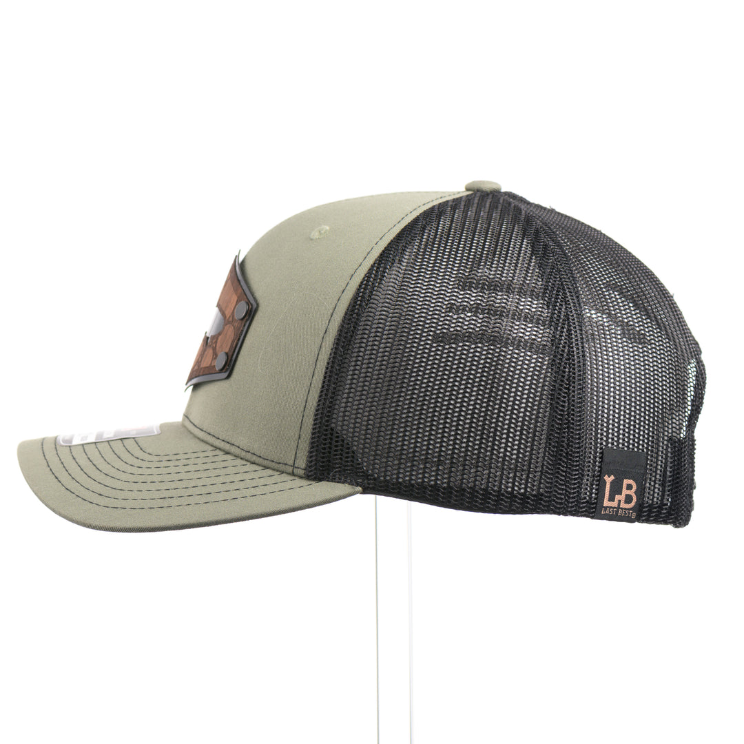 The side of a Walnut Wood Bubble Salmon Patch Trucker Hat, featuring a real walnut wood veneer patch precision-cut into a hexagon, etched with a river bubbles design with a cut-out silhouette of a salmon, laminated to a hexagonal black anodized aluminum plate, and riveted to a loden green and black mesh Richardson 112 trucker hat—handmade in Montana, USA by Last Best Supply Co.