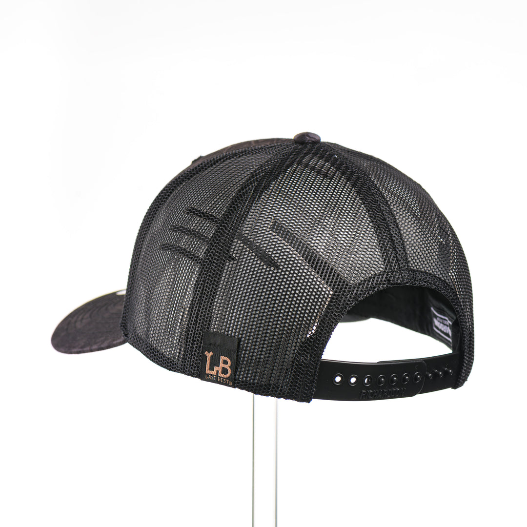 The back of a Walnut Camo Tines Kryptek Typhon Trucker Hat, featuring a hexagonal real walnut wood veneer patch with cut-out elk antler tines and laser-etched camo pattern, laminated to a hexagonal black anodized aluminum plate, and riveted to a Richardson trucker hat in Kryptek Typhon camo—handmade in Montana, USA by Last Best Supply Co.