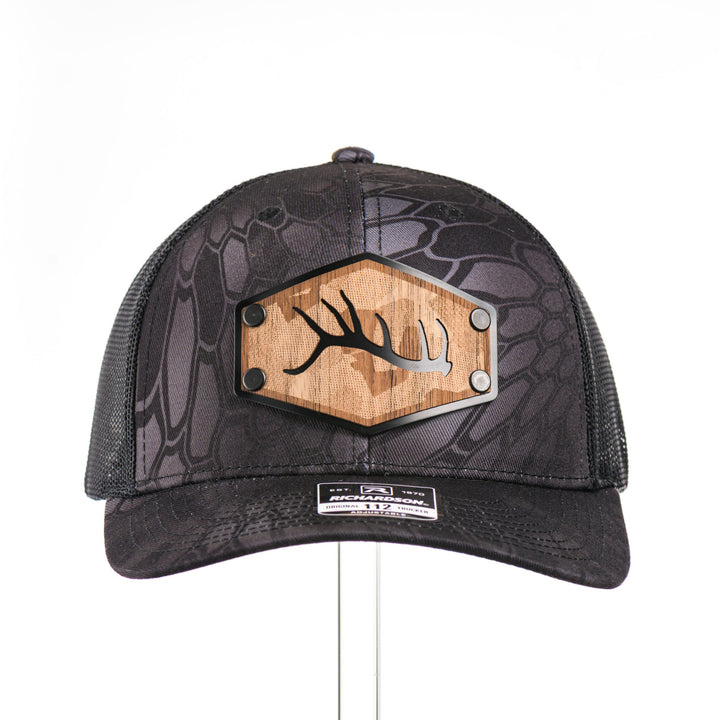 The front of a Walnut Camo Tines Kryptek Typhon Trucker Hat, featuring a hexagonal real walnut wood veneer patch with cut-out elk antler tines and laser-etched camo pattern, laminated to a hexagonal black anodized aluminum plate, and riveted to a Richardson trucker hat in Kryptek Typhon camo—handmade in Montana, USA by Last Best Supply Co.
