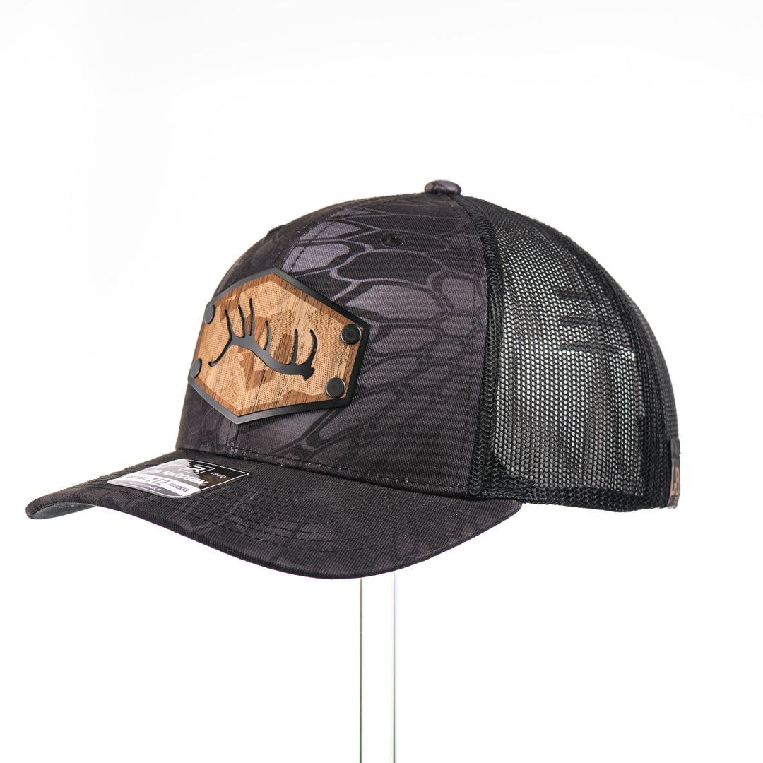 3/4 view of a Walnut Camo Tines Kryptek Typhon Trucker Hat, featuring a hexagonal real walnut wood veneer patch with cut-out elk antler tines and laser-etched camo pattern, laminated to a hexagonal black anodized aluminum plate, and riveted to a Richardson trucker hat in Kryptek Typhon camo—handmade in Montana, USA by Last Best Supply Co.