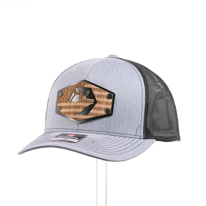 A heather gray and black Richardson trucker hat with an etched walnut wood American flag and elk patch on a hexagon metal plate at a quarter turn, handmade in Montana, USA by Last Best Supply Co.