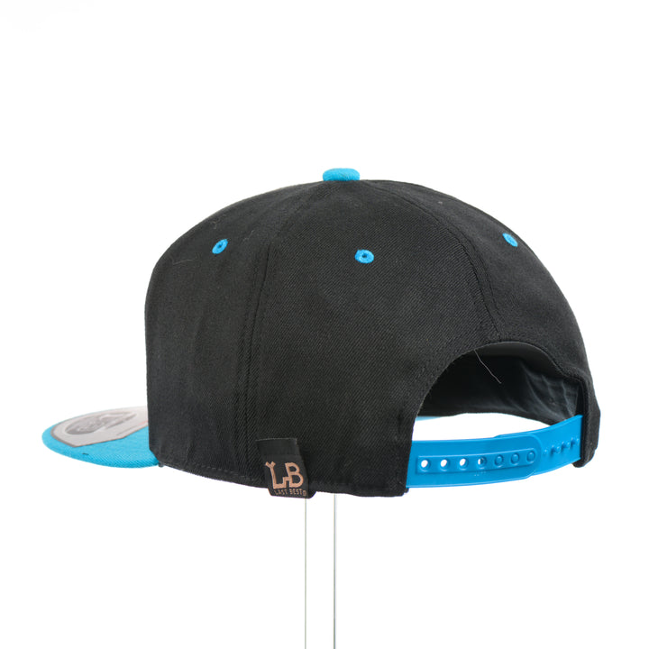 The back of a Walnut Wood Mountains Flexfit Black & Teal Flat Bill Hat, featuring a real walnut wood veneer patch etched with fine lines and cut-out mountain peaks design, laminated to a teal anodized aluminum plate and riveted to a black and teal Flexfit 110F flat bill hat—handmade in Montana, USA by Last Best Supply Co.