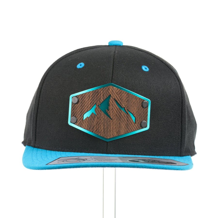 The front of a Walnut Wood Mountains Flexfit Black & Teal Flat Bill Hat, featuring a real walnut wood veneer patch etched with fine lines and cut-out mountain peaks design, laminated to a teal anodized aluminum plate and riveted to a black and teal Flexfit 110F flat bill hat—handmade in Montana, USA by Last Best Supply Co.