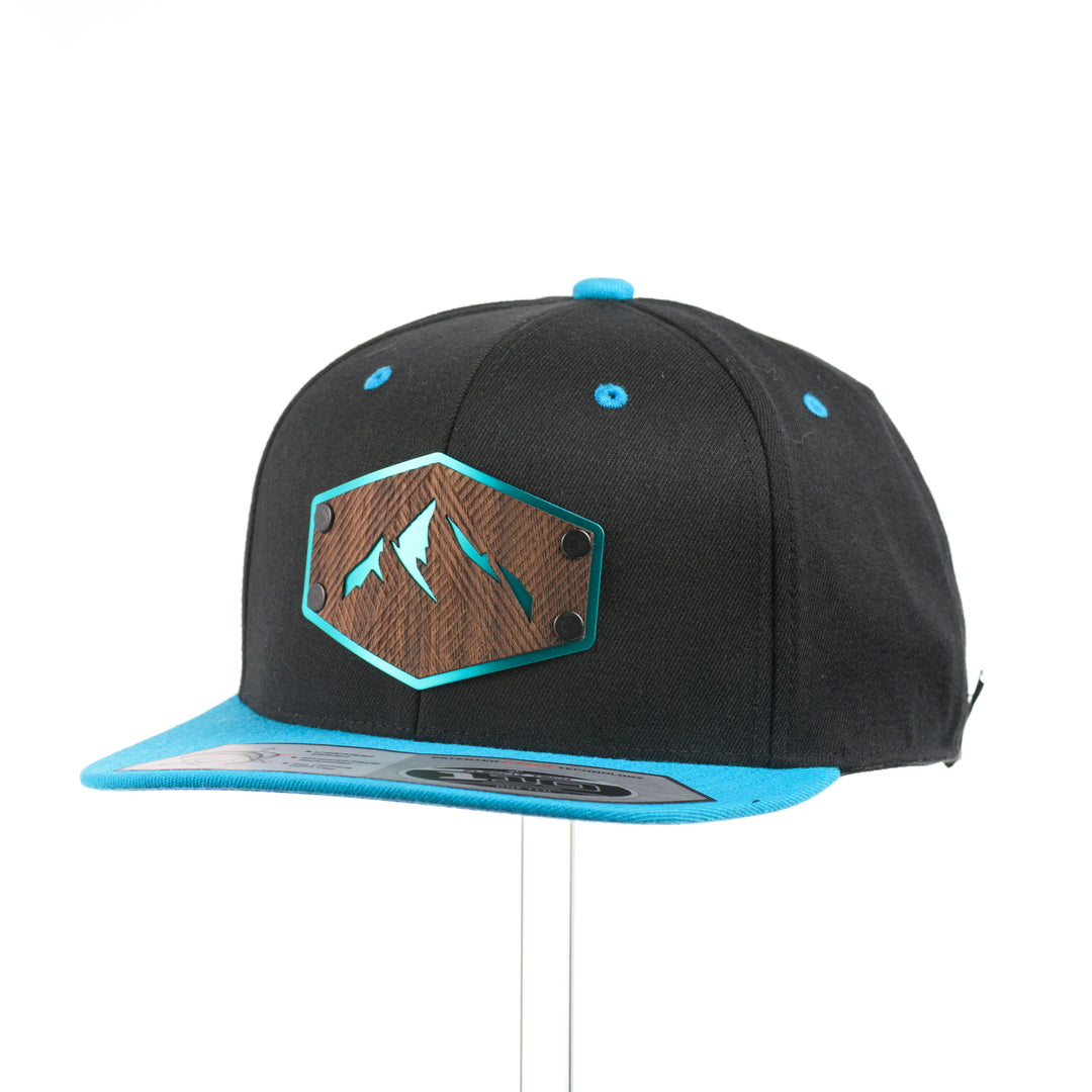 3/4 view of a Walnut Wood Mountains Flexfit Black & Teal Flat Bill Hat, featuring a real walnut wood veneer patch etched with fine lines and cut-out mountain peaks design, laminated to a teal anodized aluminum plate and riveted to a black and teal Flexfit 110F flat bill hat—handmade in Montana, USA by Last Best Supply Co.
