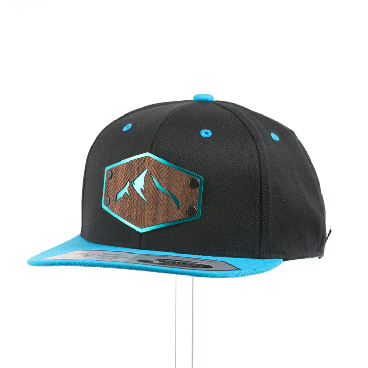 3/4 view of a Walnut Wood Mountains Flexfit Black & Teal Flat Bill Hat, featuring a real walnut wood veneer patch etched with fine lines and cut-out mountain peaks design, laminated to a teal anodized aluminum plate and riveted to a black and teal Flexfit 110F flat bill hat—handmade in Montana, USA by Last Best Supply Co.