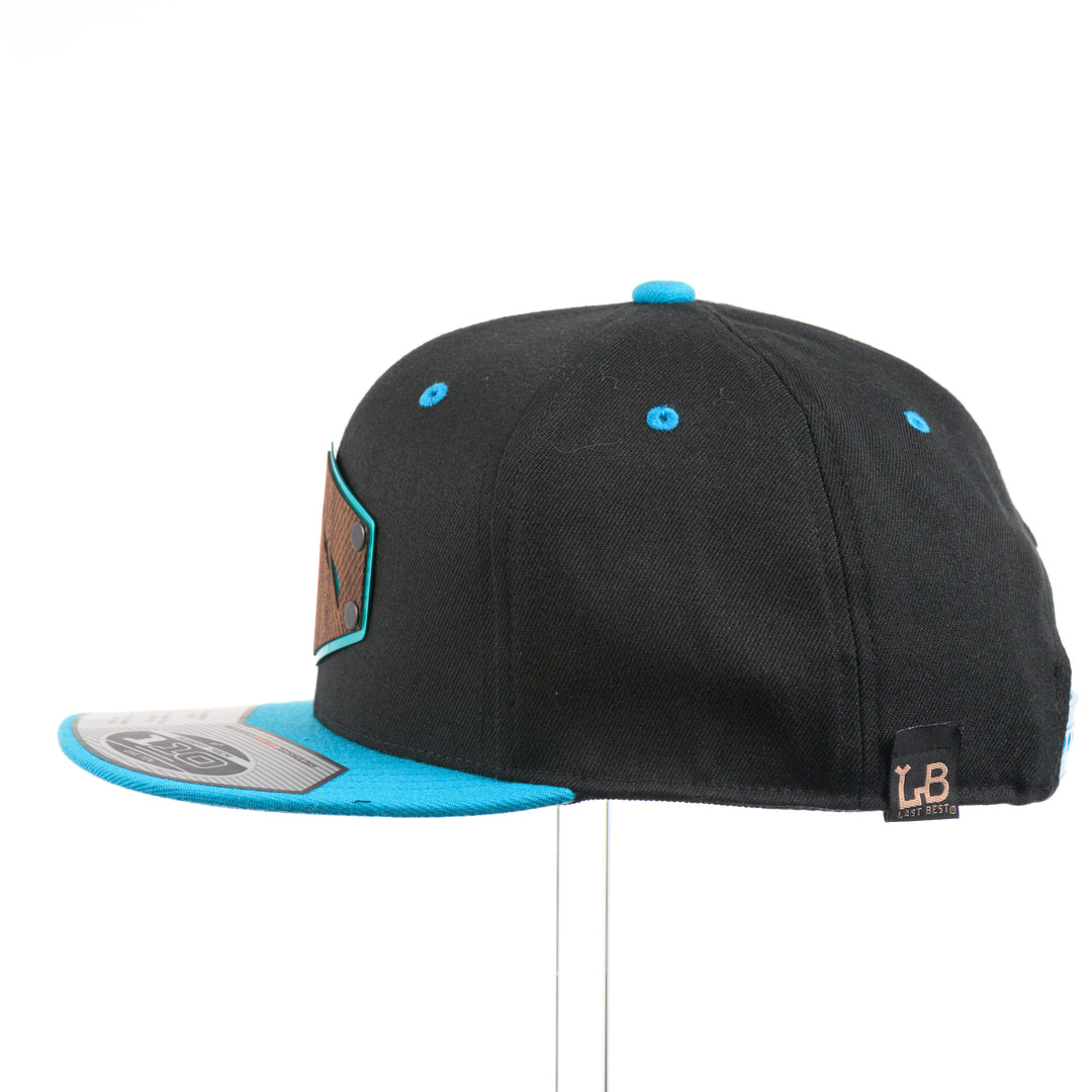 The side of a Walnut Wood Mountains Flexfit Black & Teal Flat Bill Hat, featuring a real walnut wood veneer patch etched with fine lines and cut-out mountain peaks design, laminated to a teal anodized aluminum plate and riveted to a black and teal Flexfit 110F flat bill hat—handmade in Montana, USA by Last Best Supply Co.