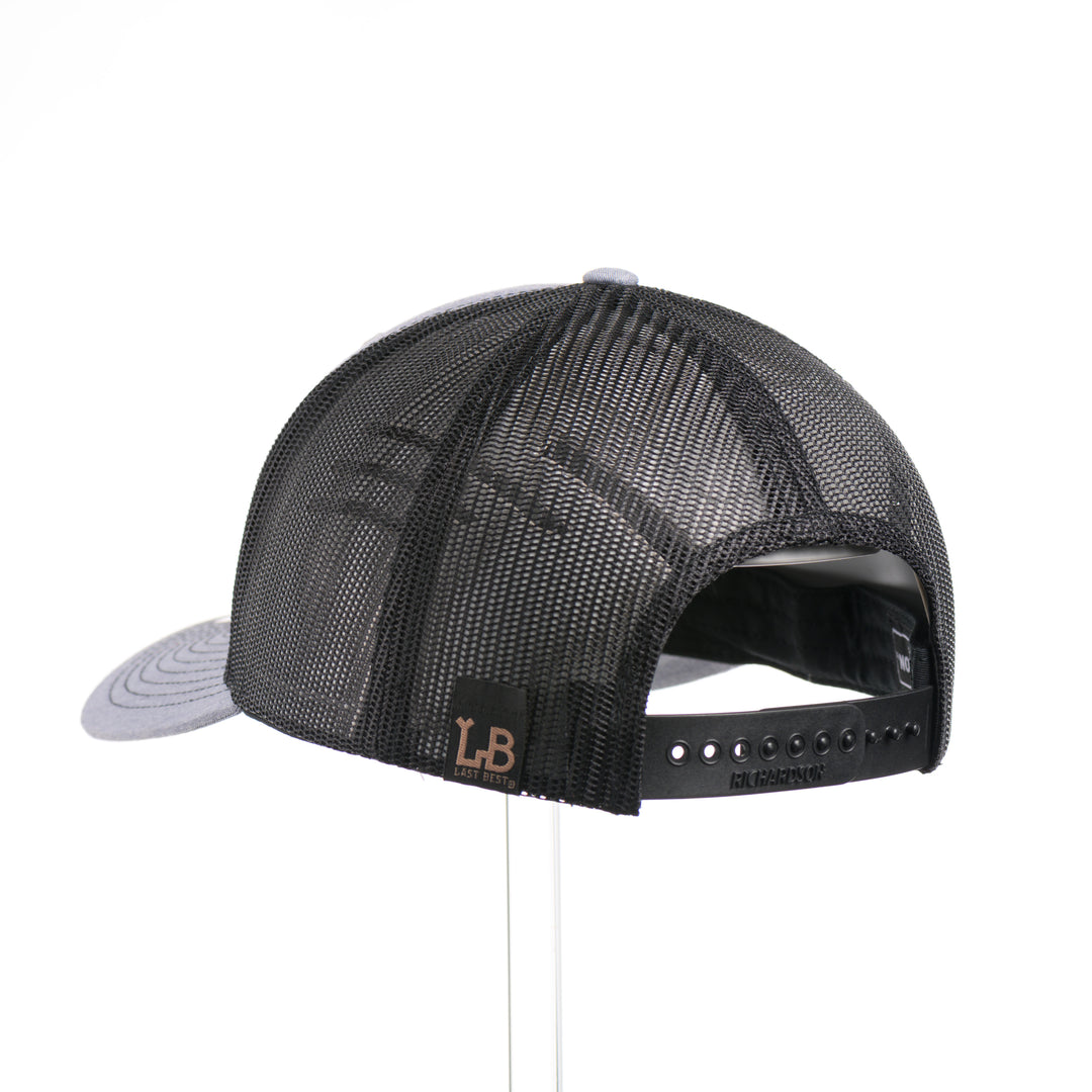 The back of a Walnut Wood Roaming Bigfoot Patch Flat Bill Trucker Hat, featuring a real walnut wood veneer patch precision-cut into a hexagon, etched with a forest scene with a silhouette of a roaming Sasquatch, laminated to a hexagonal black anodized aluminum plate, and riveted to a heather gray and black mesh Richardson 112 trucker hat—handmade in Montana, USA by Last Best Supply Co.