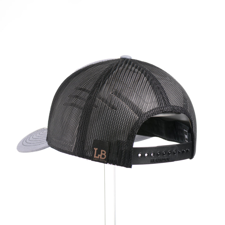 The back of a Walnut Wood Roaming Bigfoot Patch Flat Bill Trucker Hat, featuring a real walnut wood veneer patch precision-cut into a hexagon, etched with a forest scene with a silhouette of a roaming Sasquatch, laminated to a hexagonal black anodized aluminum plate, and riveted to a heather gray and black mesh Richardson 112 trucker hat—handmade in Montana, USA by Last Best Supply Co.