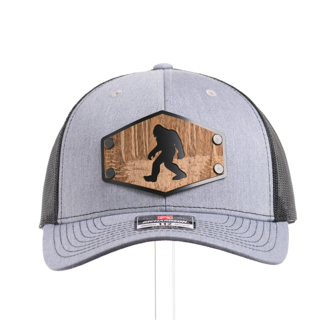 The front of a Walnut Wood Roaming Bigfoot Patch Flat Bill Trucker Hat, featuring a real walnut wood veneer patch precision-cut into a hexagon, etched with a forest scene with a silhouette of a roaming Sasquatch, laminated to a hexagonal black anodized aluminum plate, and riveted to a heather gray and black mesh Richardson 112 trucker hat—handmade in Montana, USA by Last Best Supply Co.