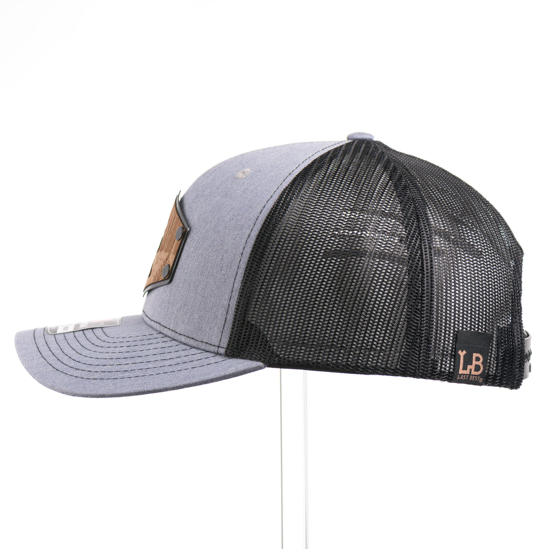 The side of a Walnut Wood Roaming Bigfoot Patch Flat Bill Trucker Hat, featuring a real walnut wood veneer patch precision-cut into a hexagon, etched with a forest scene with a silhouette of a roaming Sasquatch, laminated to a hexagonal black anodized aluminum plate, and riveted to a heather gray and black mesh Richardson 112 trucker hat—handmade in Montana, USA by Last Best Supply Co.