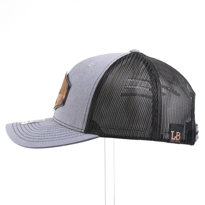 The side of a Walnut Wood Roaming Bigfoot Patch Flat Bill Trucker Hat, featuring a real walnut wood veneer patch precision-cut into a hexagon, etched with a forest scene with a silhouette of a roaming Sasquatch, laminated to a hexagonal black anodized aluminum plate, and riveted to a heather gray and black mesh Richardson 112 trucker hat—handmade in Montana, USA by Last Best Supply Co.