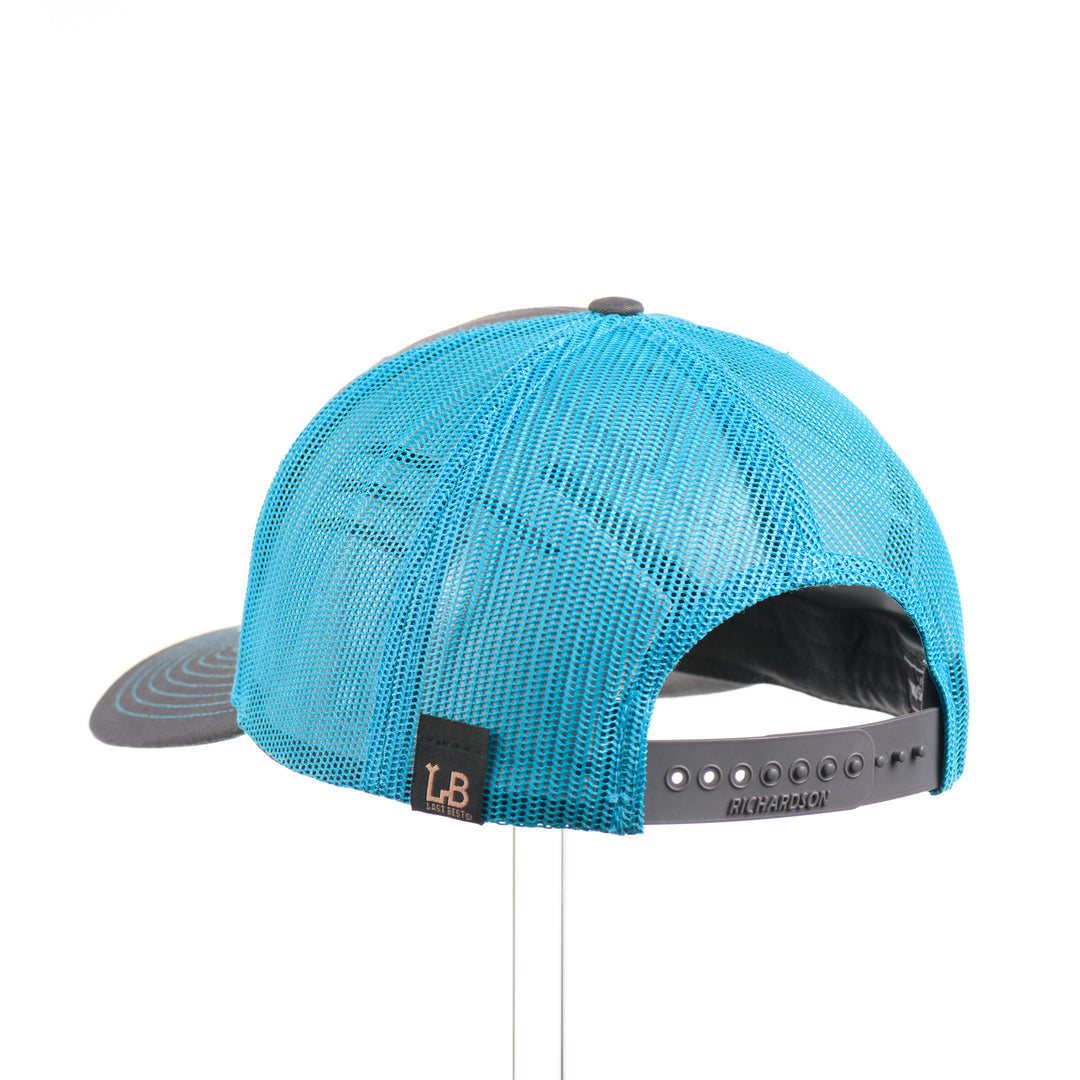 The back of a bright teal and charcoal Richardson trucker hat with an etched walnut patch featuring trees and cut out lines on a teal hexagon-shaped anodized aluminum plate—handmade in Montana, USA by Last Best Supply Co.