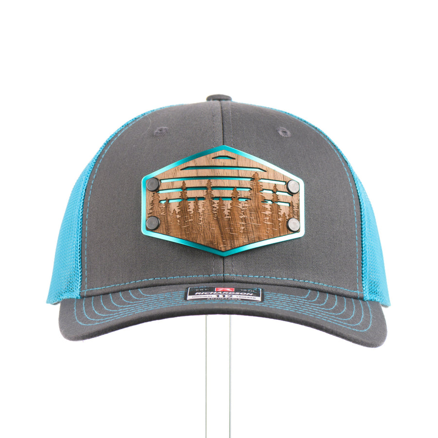 The front of a bright teal and charcoal Richardson trucker hat with an etched walnut patch featuring trees and cut out lines on a teal hexagon-shaped anodized aluminum plate—handmade in Montana, USA by Last Best Supply Co.