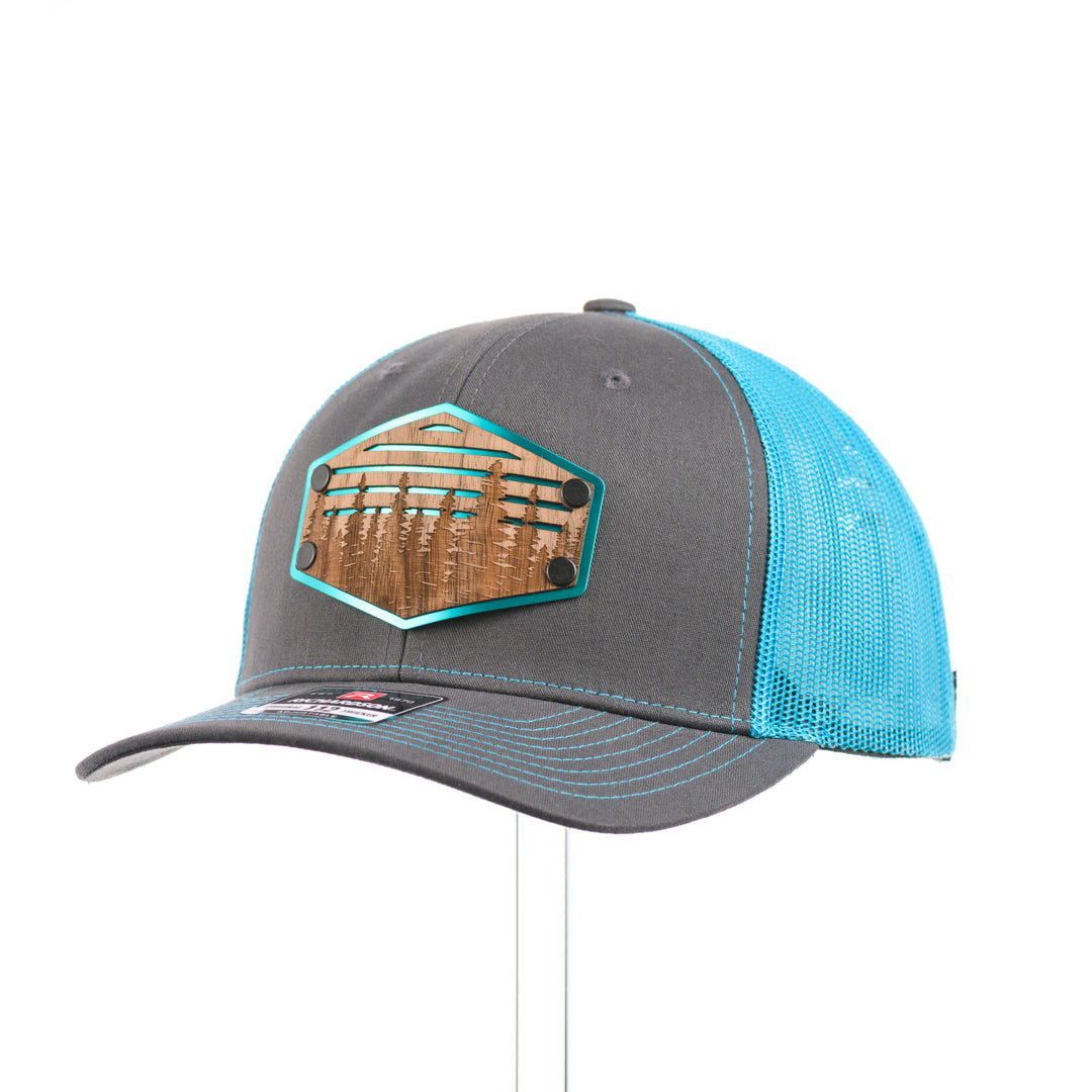 A bright teal and charcoal Richardson trucker hat with an etched walnut patch featuring trees and cut out lines on a teal hexagon-shaped anodized aluminum plate—handmade in Montana, USA by Last Best Supply Co.--at a quarter turn.