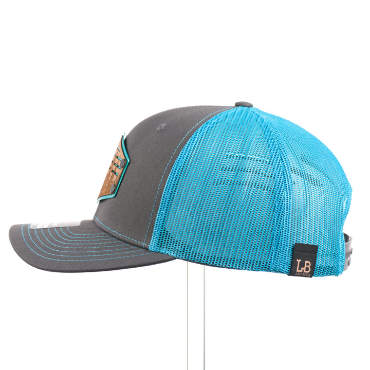 The side of a bright teal and charcoal Richardson trucker hat with an etched walnut patch featuring trees and cut out lines on a teal hexagon-shaped anodized aluminum plate—handmade in Montana, USA by Last Best Supply Co.