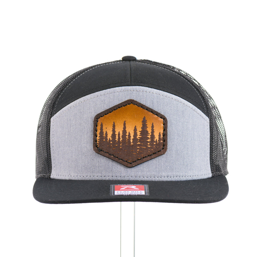 The front of an Evergreen Trees Hexagon Leather Patch Flat Bill Trucker Hat, featuring a hexagonal hand-dyed leather patch etched with an evergreen forest scene, securely sewn to the structured front of a heather gray and black Richardson 168 seven-panel flat bill trucker hat—handmade in Montana, USA by Last Best Supply Co.