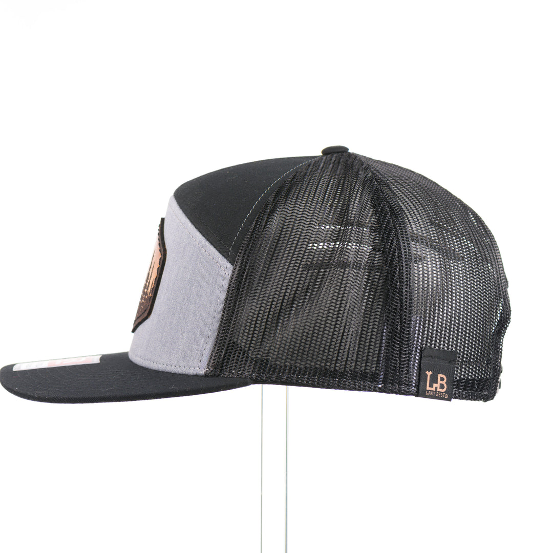 The side of an Evergreen Trees Hexagon Leather Patch Flat Bill Trucker Hat, featuring a hexagonal hand-dyed leather patch etched with an evergreen forest scene, securely sewn to the structured front of a heather gray and black Richardson 168 seven-panel flat bill trucker hat—handmade in Montana, USA by Last Best Supply Co.