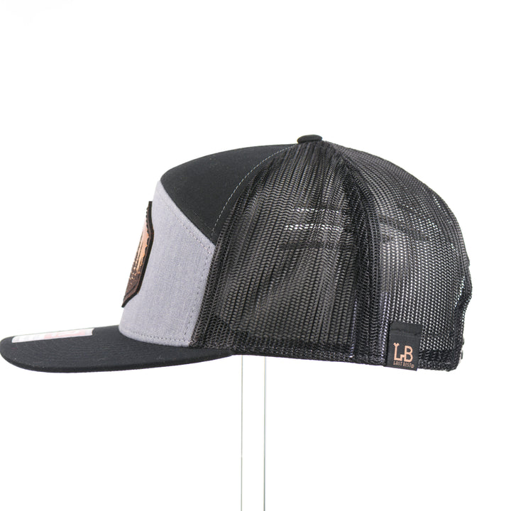The side of an Evergreen Trees Hexagon Leather Patch Flat Bill Trucker Hat, featuring a hexagonal hand-dyed leather patch etched with an evergreen forest scene, securely sewn to the structured front of a heather gray and black Richardson 168 seven-panel flat bill trucker hat—handmade in Montana, USA by Last Best Supply Co.