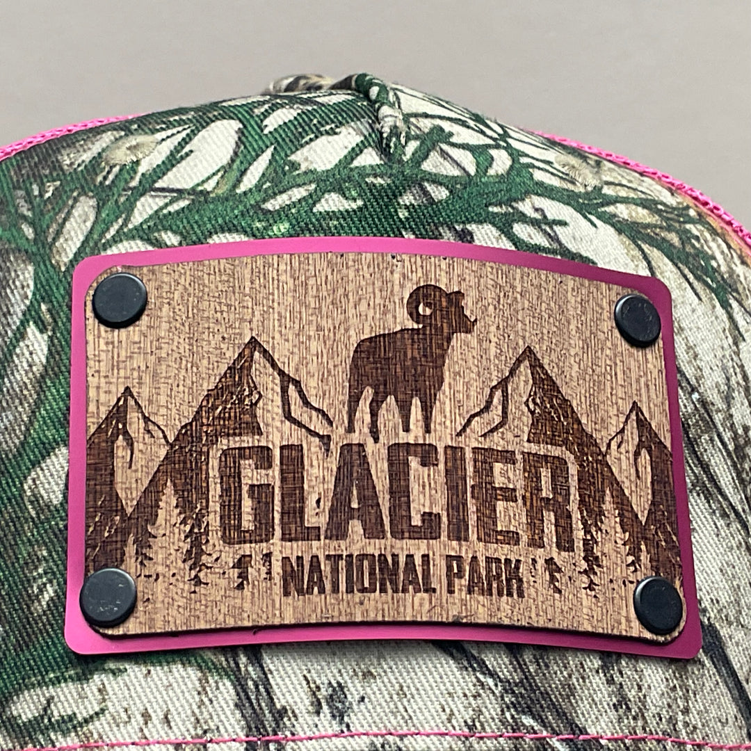 Patch detail of an Explore Glacier National Park Wood Patch on Real Tree Pink Camo Richardson Hat, featuring a rectangular, real wood veneer patch etched with Glacier National Park and a bighorn sheep, laminated to a pink anodized metal plate, and then riveted to an Real Tree camo and pink Richardson 112 trucker hat—handmade in Montana, USA by Last Best Supply Co.