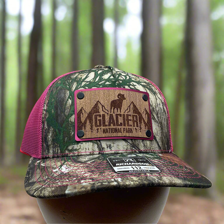 Front of an Explore Glacier National Park Wood Patch on Real Tree Pink Camo Richardson Hat, featuring a rectangular, real wood veneer patch etched with Glacier National Park and a bighorn sheep, laminated to a pink anodized metal plate, and then riveted to an Real Tree camo and pink Richardson 112 trucker hat—handmade in Montana, USA by Last Best Supply Co.