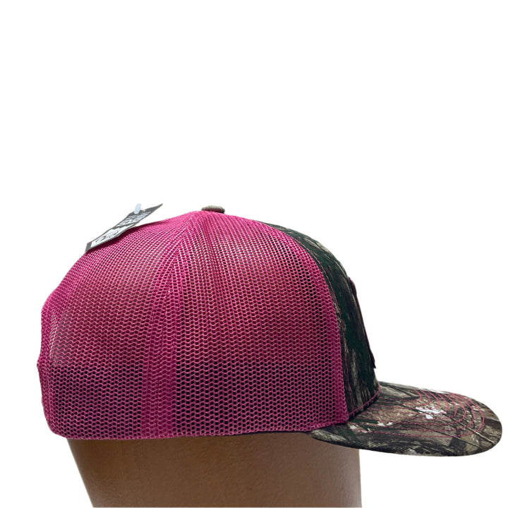 Side view of a Explore Glacier National Park Wood Patch on Real Tree Pink Camo Richardson Hat, featuring a rectangular, real wood veneer patch etched with Glacier National Park and a bighorn sheep, laminated to a pink anodized metal plate, and then riveted to an Real Tree camo and pink Richardson 112 trucker hat—handmade in Montana, USA by Last Best Supply Co.