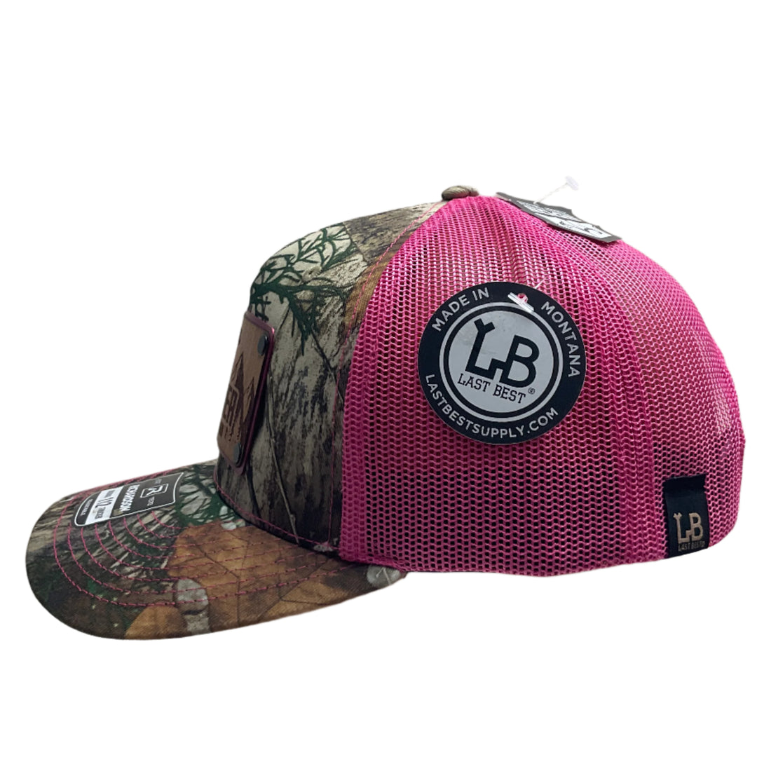 Side view of an Explore Glacier National Park Wood Patch on Real Tree Pink Camo Richardson Hat, featuring a rectangular, real wood veneer patch etched with Glacier National Park and a bighorn sheep, laminated to a pink anodized metal plate, and then riveted to an Real Tree camo and pink Richardson 112 trucker hat—handmade in Montana, USA by Last Best Supply Co.