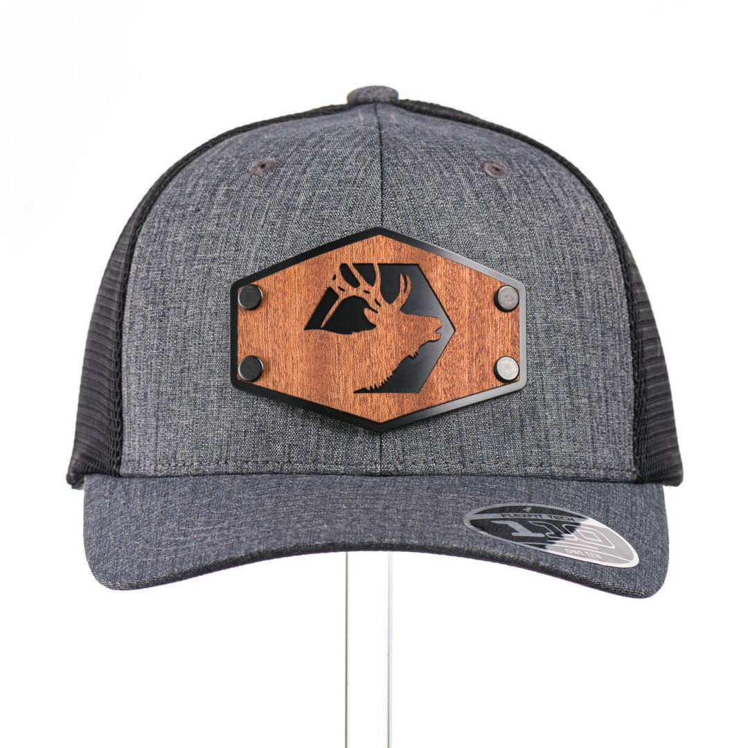 The front of a Mahogany Wood Elk Patch Flexfit Trucker Hat, featuring a real mahogany wood veneer patch precision-cut into a hexagon shape with a bold elk silhouette cut-out, laminated to a matching anodized aluminum plate, and riveted to a charcoal and black Flexfit 110F flat bill hat—handmade in Montana, USA by Last Best Supply Co.