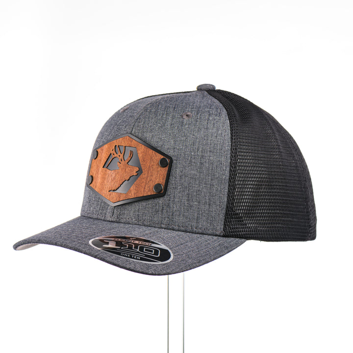 3/4 view of a Mahogany Wood Elk Patch Flexfit Trucker Hat, featuring a real mahogany wood veneer patch precision-cut into a hexagon shape with a bold elk silhouette cut-out, laminated to a matching anodized aluminum plate, and riveted to a charcoal and black Flexfit 110F flat bill hat—handmade in Montana, USA by Last Best Supply Co.