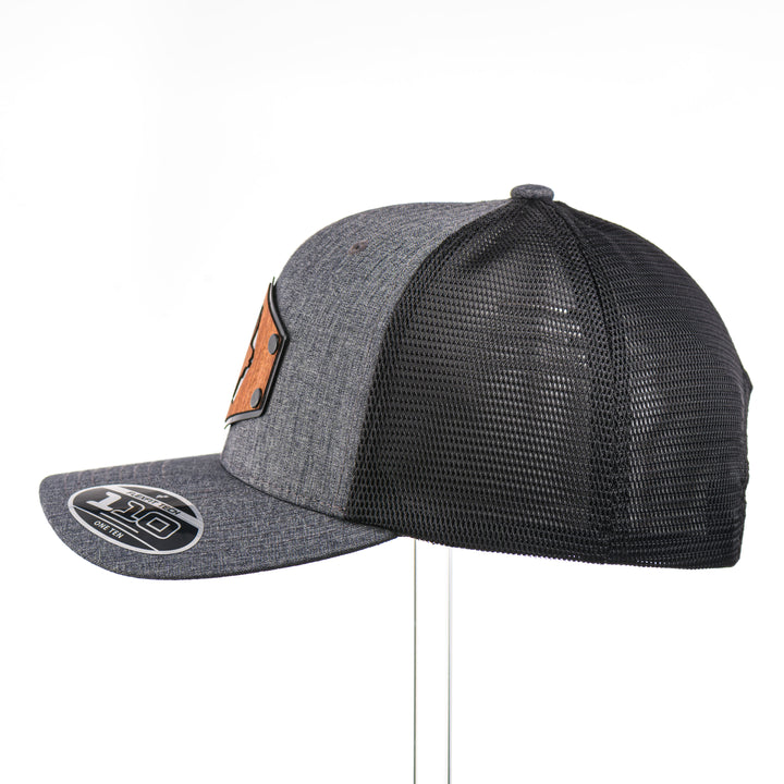 The side view of a Mahogany Wood Elk Patch Flexfit Trucker Hat, featuring a real mahogany wood veneer patch precision-cut into a hexagon shape with a bold elk silhouette cut-out, laminated to a matching anodized aluminum plate, and riveted to a charcoal and black Flexfit 110F flat bill hat—handmade in Montana, USA by Last Best Supply Co.