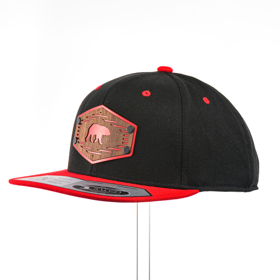 3/4 view of a Grizzly Bear w/ Lines Walnut Wood Black & Red Flat Bill Hat, featuring a hexagonal real walnut wood veneer patch with a cut-out bear silhouette and fine line details, laminated to a hexagonal red anodized aluminum plate, and riveted to a black and red Flexfit 110F flat bill hat—handmade in Montana, USA by Last Best Supply Co.